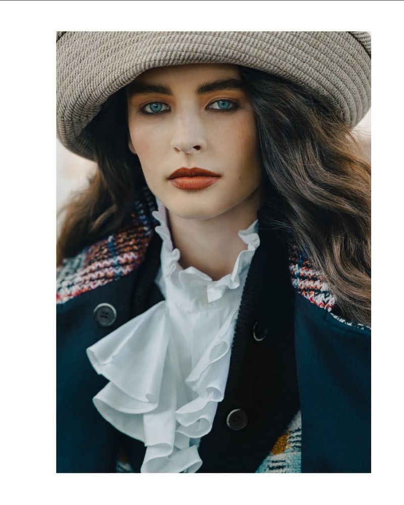 A woman with blue eyes and wavy hair is wearing a beige hat, a white ruffled shirt, and a dark coat in a close-up studio shot.