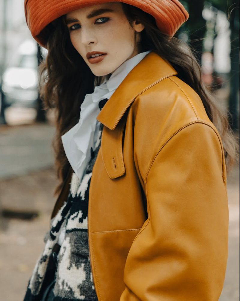 Stylish woman in an orange hat and leather jacket posing outdoors, showcasing a fashionable editorial portrait with a blurry background.