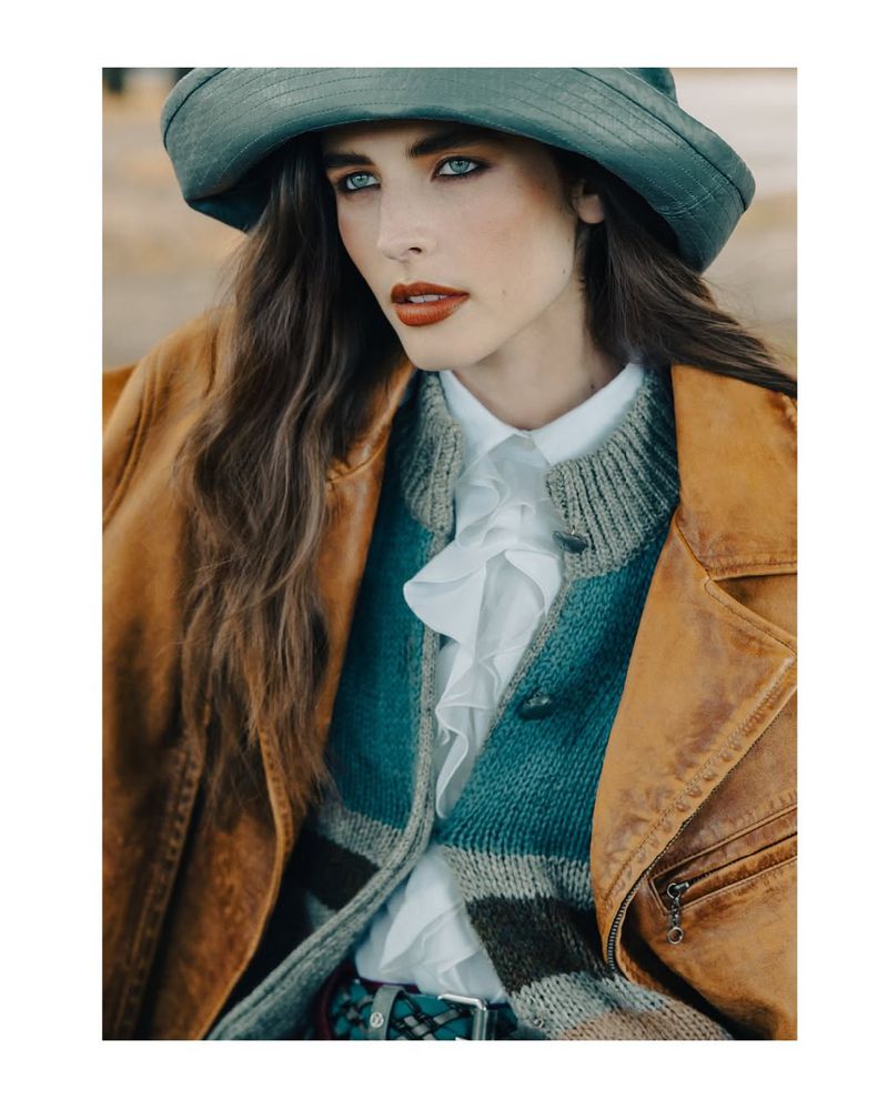 Portrait of a woman wearing a hat and a brown jacket. She has blue eyes and red lipstick, looking directly at the camera.