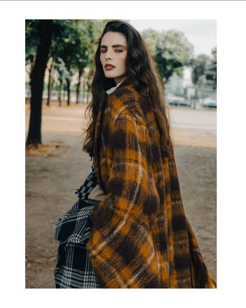 Portrait of a woman with long brown hair wearing a plaid coat in an outdoor setting with trees in the background.