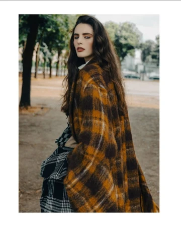 Portrait of a woman with long brown hair wearing a plaid coat in an outdoor setting with trees in the background.