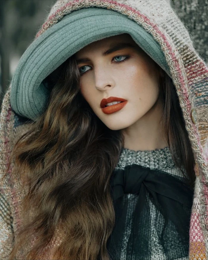Close-up portrait of a model wearing a green hat and hooded tweed coat with bold rust-red lipstick and wavy dark hair.