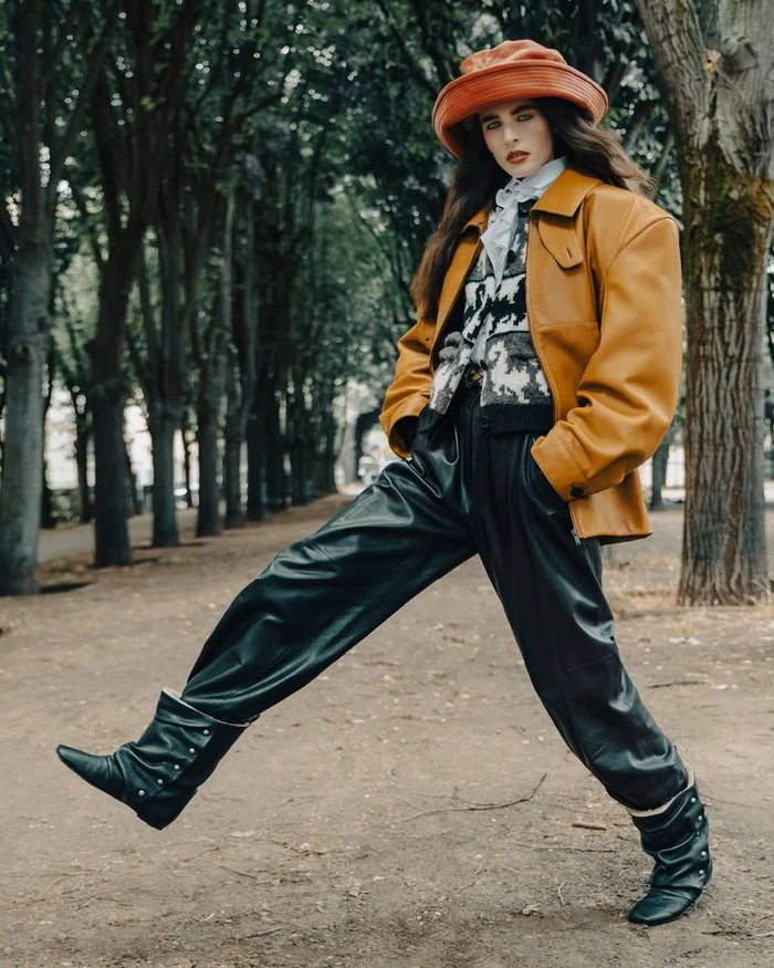 Model wearing a tan leather jacket, patterned sweater, bucket hat, and black pants stepping across a park path lined with trees.