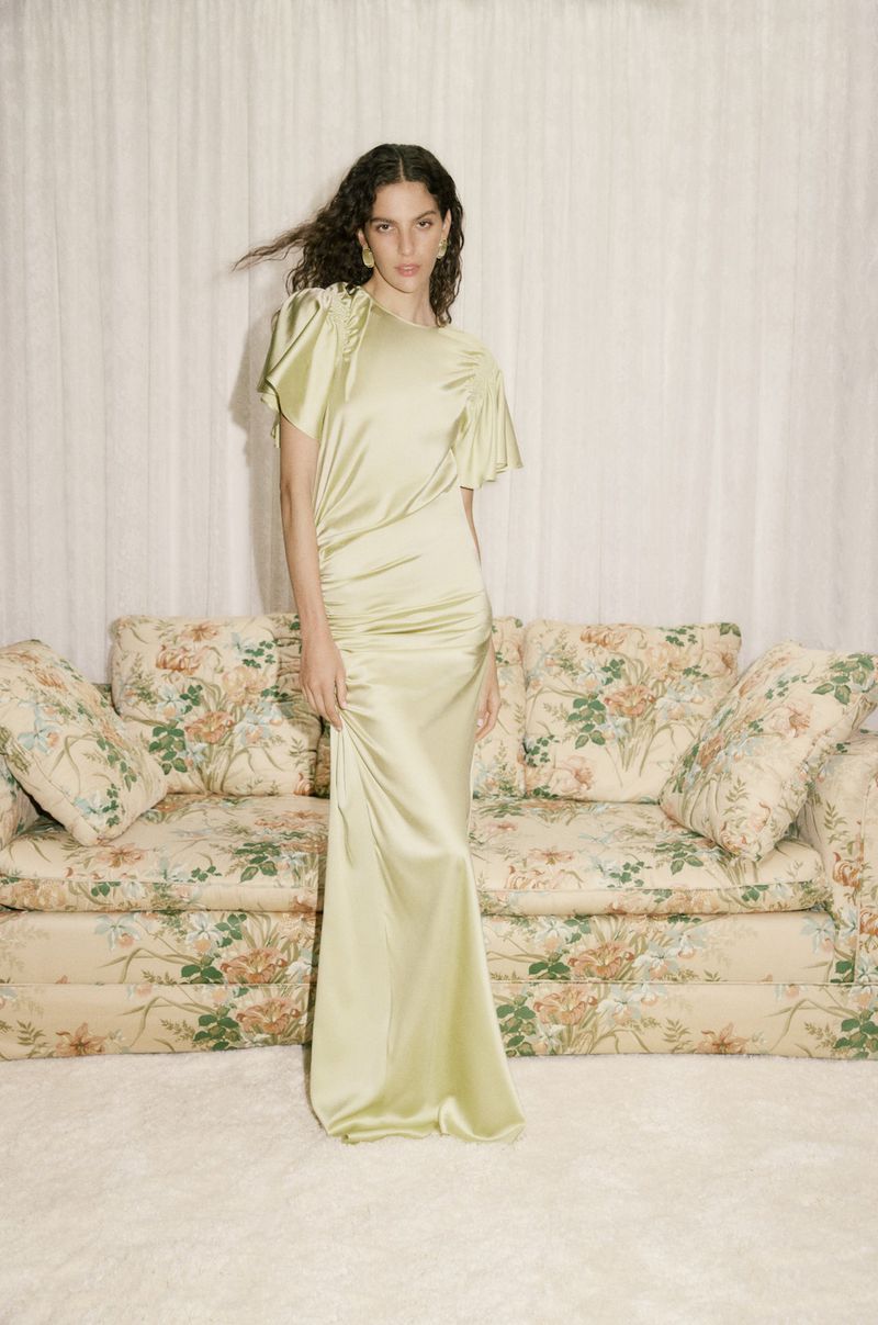 A woman poses indoors in a pale green formal gown, standing in front of a floral couch and cream-colored curtain.