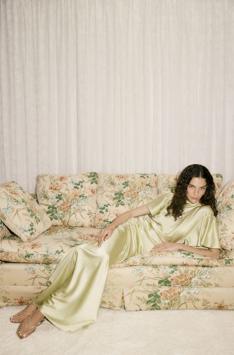 A woman lounges in a light green dress on a floral couch, posing in an indoor setting with a soft, neutral color palette.