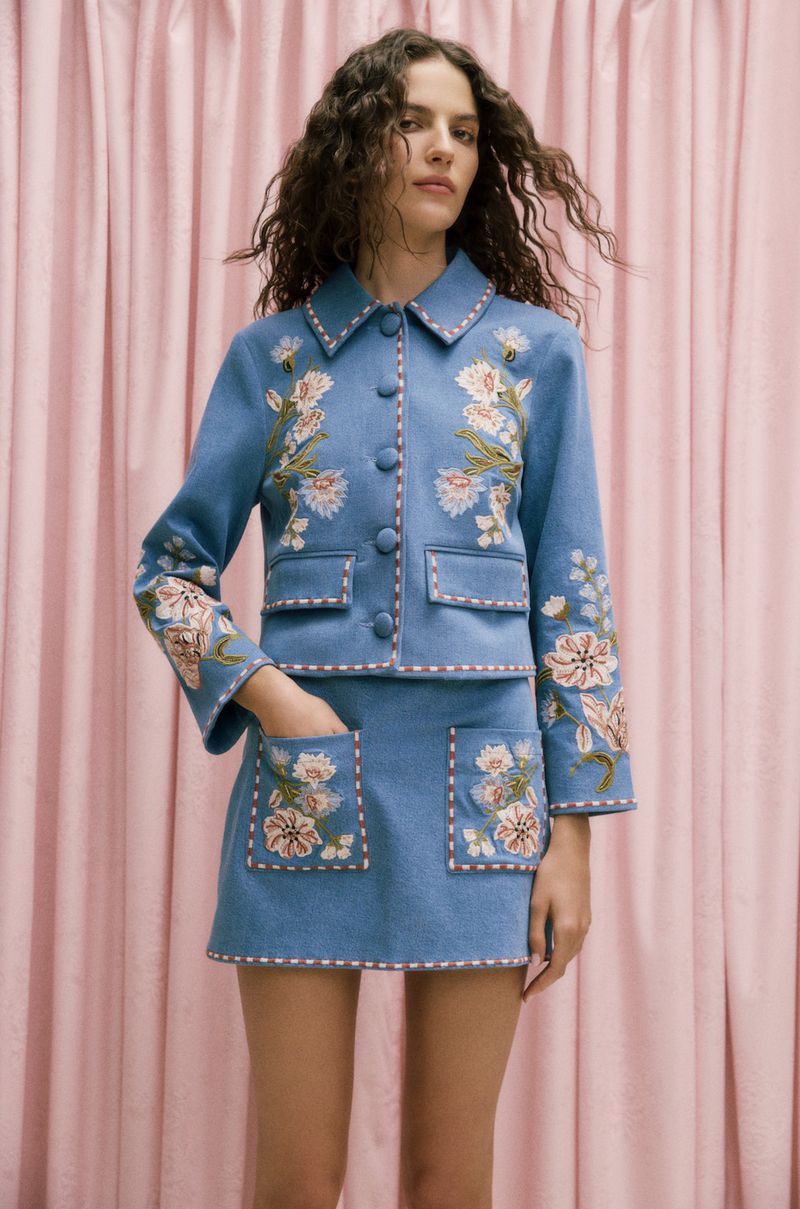 A woman in a light blue embroidered jacket and skirt poses against a pink curtain, showcasing a spring fashion style.