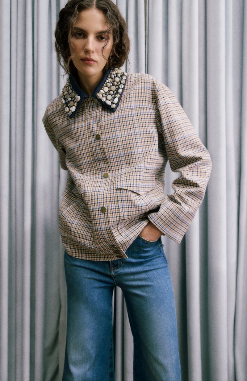 Woman in a plaid jacket with pearl collar and blue jeans, posing against a curtain. Fashion shot with natural lighting.