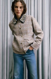 Model wearing a plaid button-down jacket with an embellished collar and blue denim jeans against grey pleated curtains.