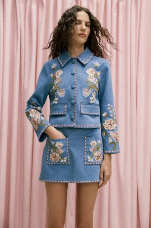 Model Africa Garcia poses in a light blue embroidered jacket and matching skirt against a draped pink fabric curtain background.