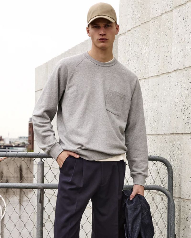 Fashion shot of a young male model wearing a gray sweatshirt and navy pants, posing in front of a textured wall and railing.