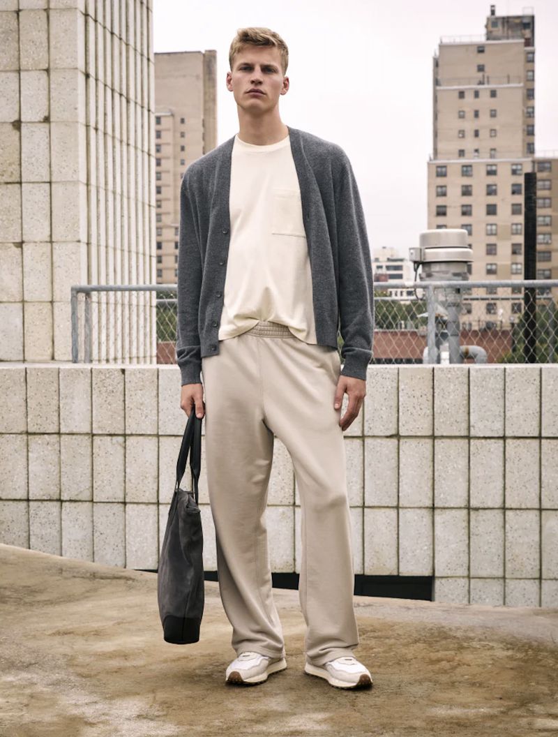 Adult male poses in a relaxed fashion-style shoot, outdoors on a rooftop with city buildings in the background, wearing casual attire.