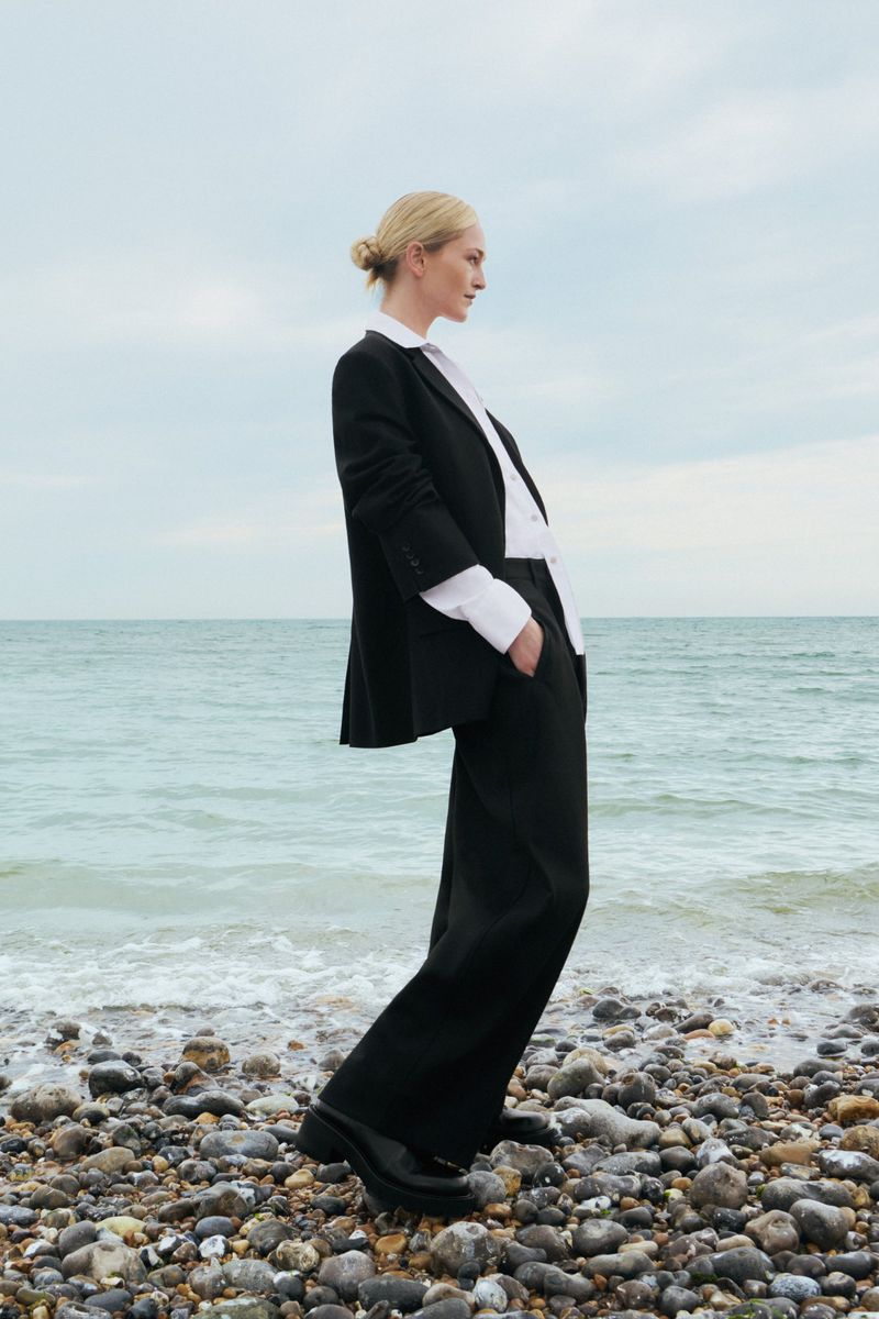 Elegant blonde woman in a black blazer and trousers stands on a rocky beach with waves gently lapping, under an overcast sky.
