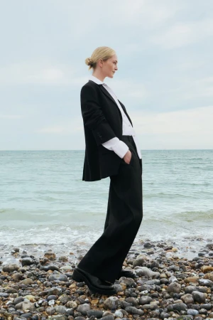 Elegant blonde woman in a black blazer and trousers stands on a rocky beach with waves gently lapping, under an overcast sky.