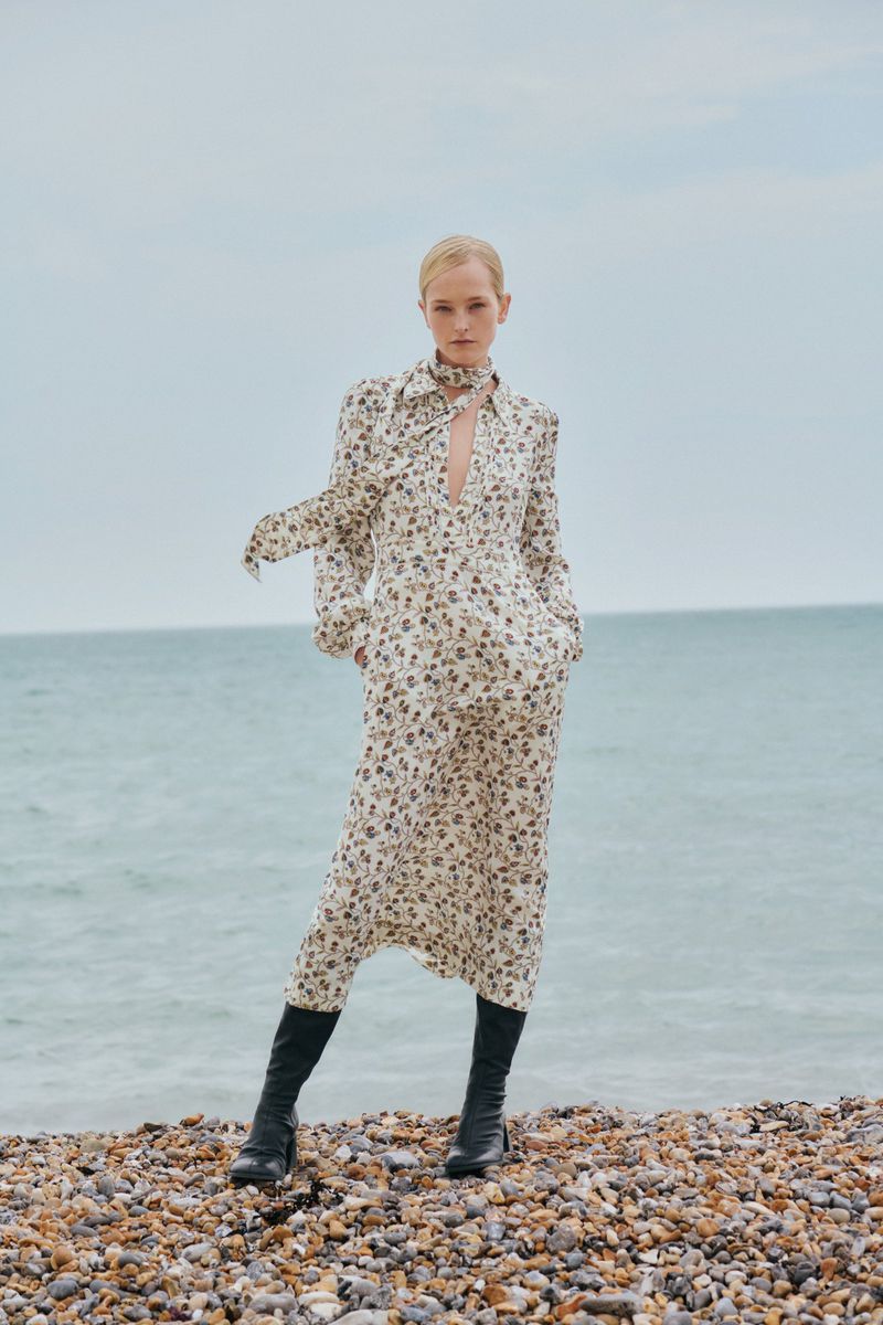 A fair-skinned woman poses in a floral dress and black boots on a rocky beach with a backdrop of sea and overcast sky.