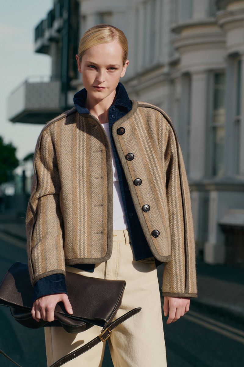Adult female wearing a striped jacket and trousers, holding a dark brown leather clutch with a shoulder strap in an outdoor urban setting.