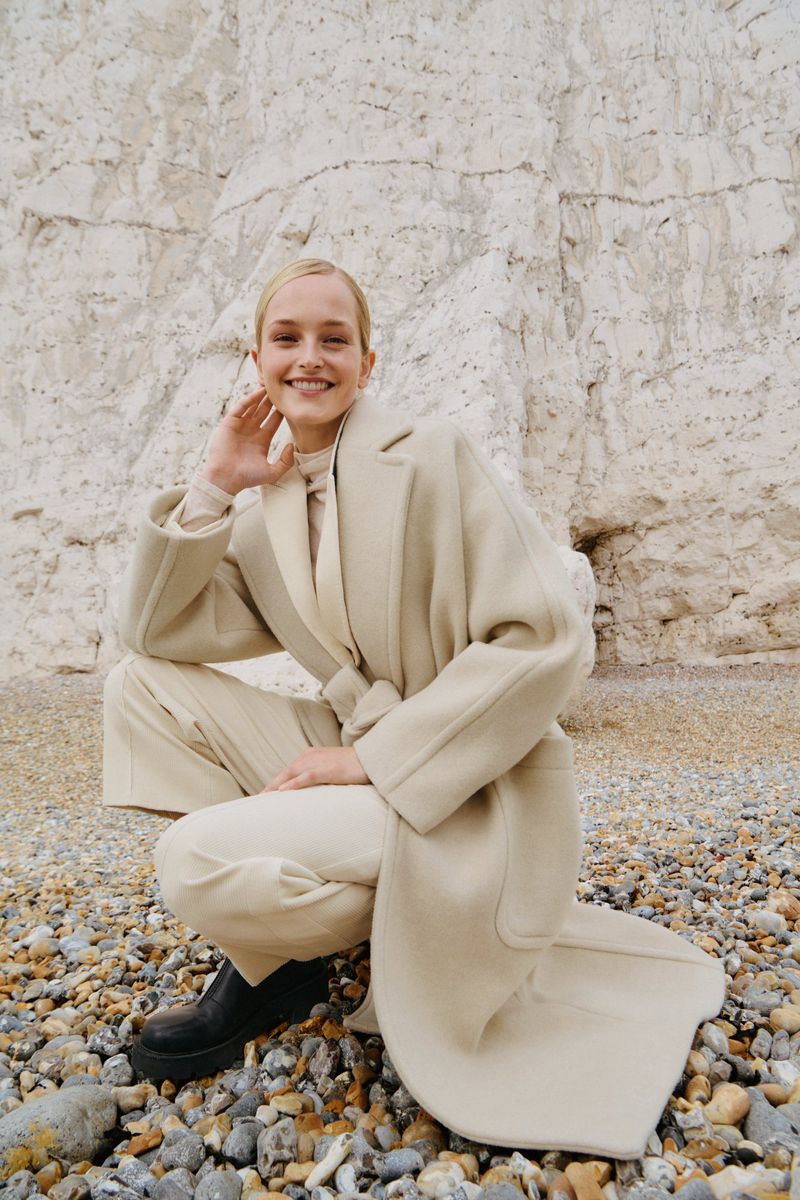 Blond woman in a beige coat poses smiling on a pebble beach against a light stone cliff backdrop for a fashion editorial.