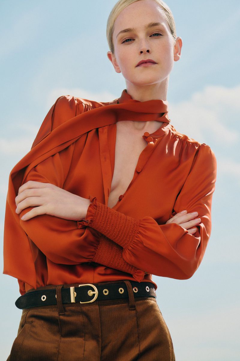 Blond woman in an orange blouse and brown pants poses against a blue sky background in a fashion or editorial style photo.