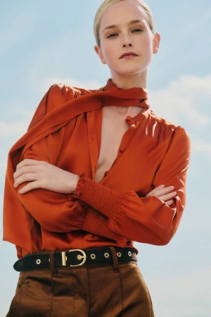 Blond woman in an orange blouse and brown pants poses against a blue sky background in a fashion or editorial style photo.