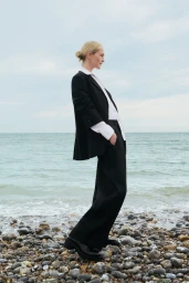 Model Jean Campbell wearing a black tailored suit and white collared shirt standing on a pebble beach by the sea.