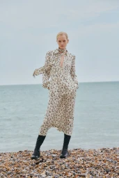 Model Jean Campbell wearing a floral print midi dress and black knee-high boots on a pebble beach overlooking the ocean.
