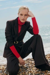 Model Jean Campbell wearing a dark pinstripe suit and red turtleneck, crouching on a pebble beach by the sea.