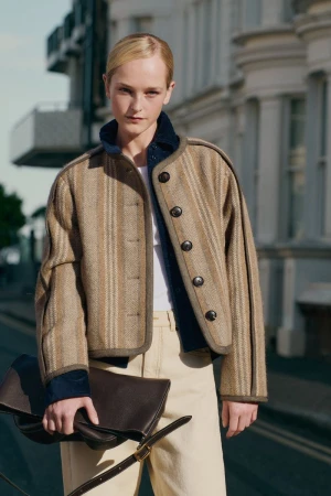 Model Jean Campbell wearing a textured striped cropped jacket and cream trousers outdoors with a dark brown leather clutch bag.