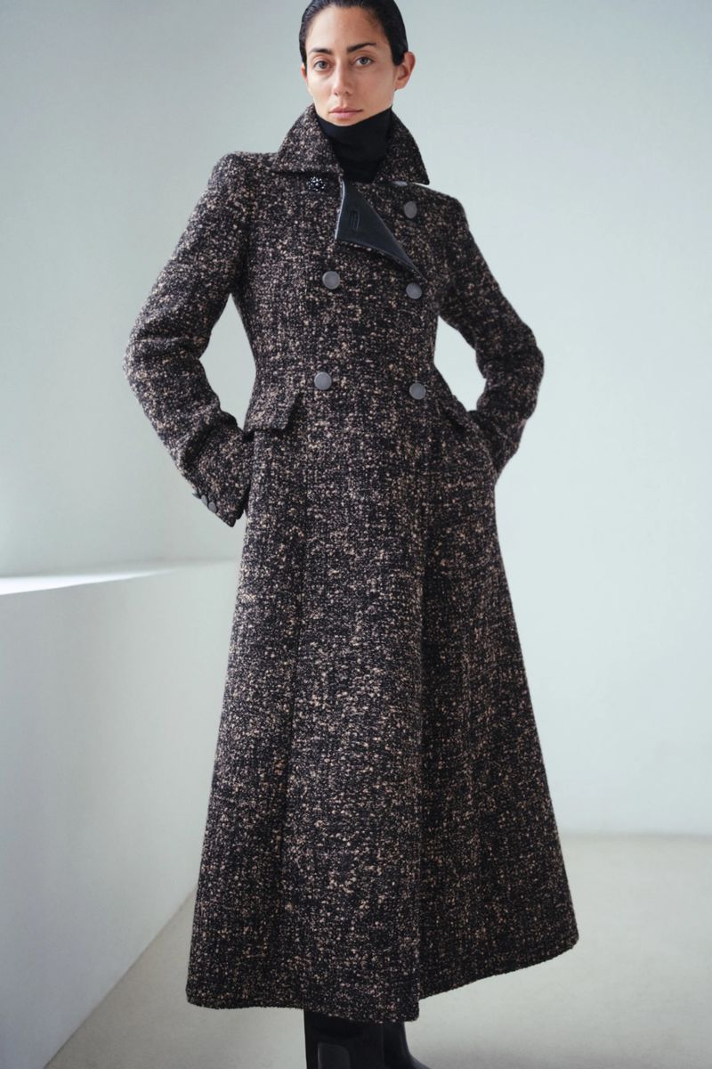 A fashionable adult female stands indoors wearing a long, patterned tweed coat, black turtleneck, and black boots against a plain backdrop.