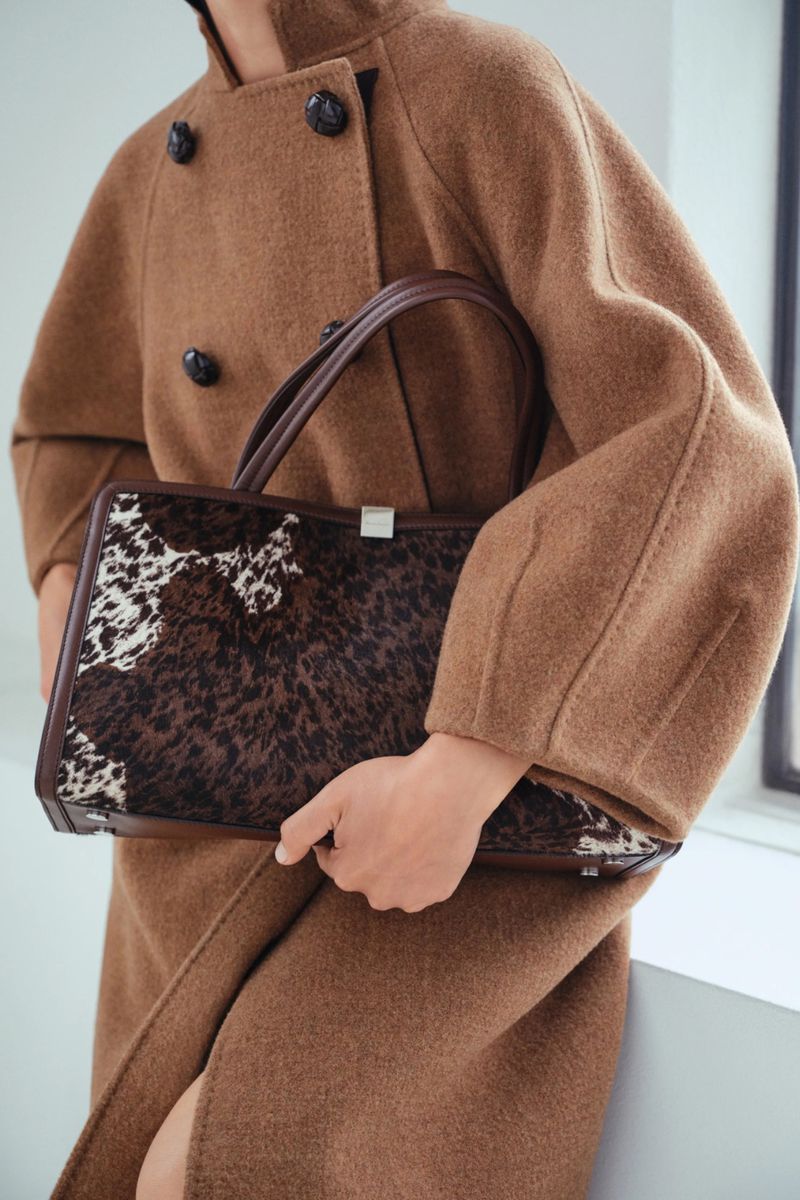 Adult female wearing a brown wool coat holds a brown and white animal-print handbag near a bright window in a studio setting.