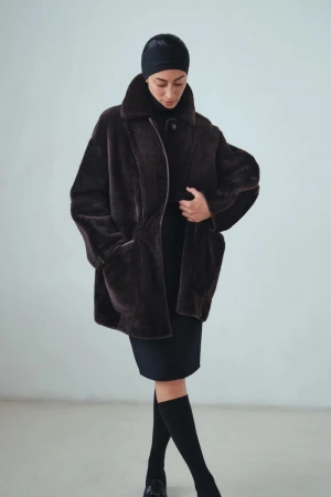 Model wearing an oversized dark brown plush shearling coat with a black head cap and skirt against a pale studio background.
