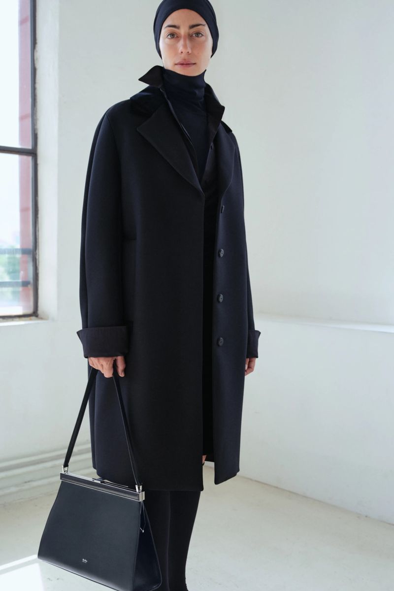 Fashion shot of an adult woman in a long black coat, black beanie, and black purse, standing in a bright, minimalist interior.