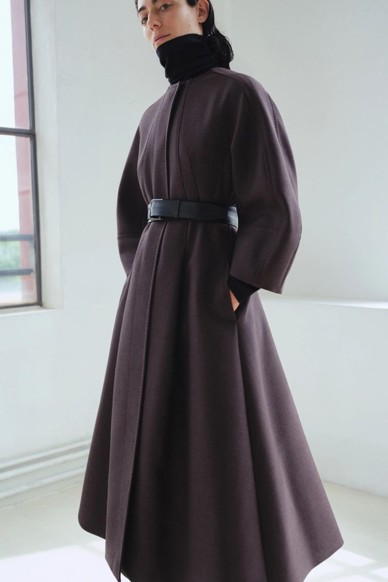 Woman in a dark brown, belted wool coat stands indoors near a bright window with natural light. Fashion shot. Fall/Winter collection.