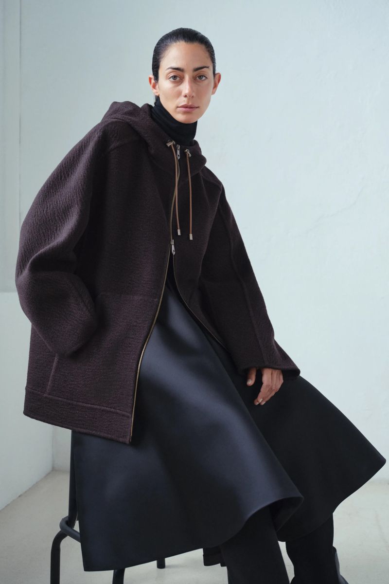 A fashionable woman is sitting in a studio, wearing a brown hooded jacket, and black dress, against a light wall.