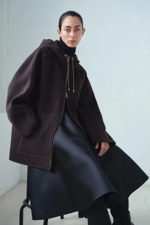 A fashionable woman is sitting in a studio, wearing a brown hooded jacket, and black dress, against a light wall.