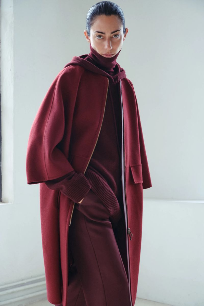 Woman poses in a hooded turtleneck sweater and matching long coat in a minimalist setting, showcasing fall fashion.
