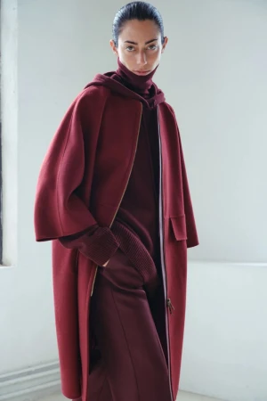 Woman poses in a hooded turtleneck sweater and matching long coat in a minimalist setting, showcasing fall fashion.