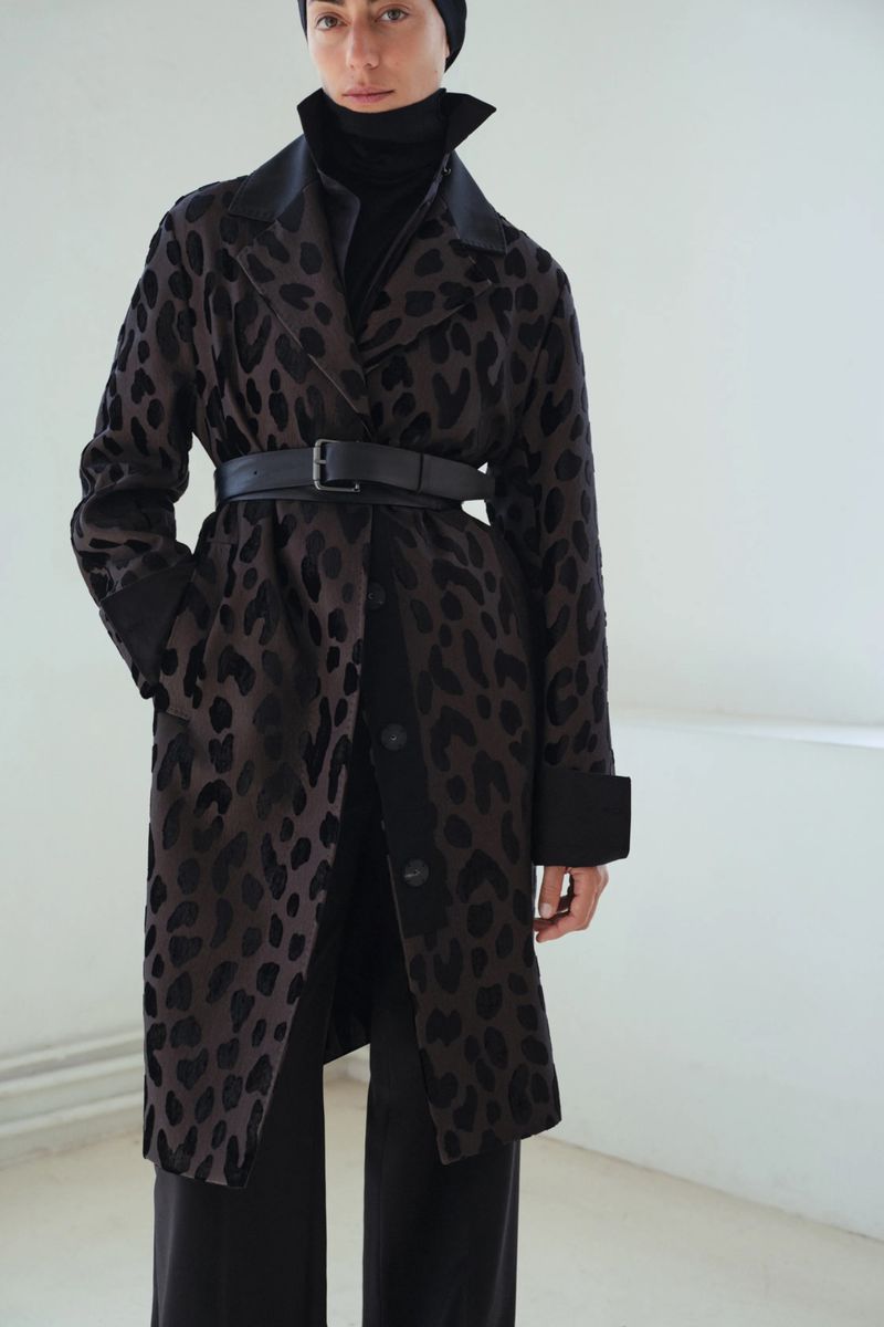 A woman wearing a long leopard print coat and a black beanie stands indoors against a white wall, posing for a fashion shot.