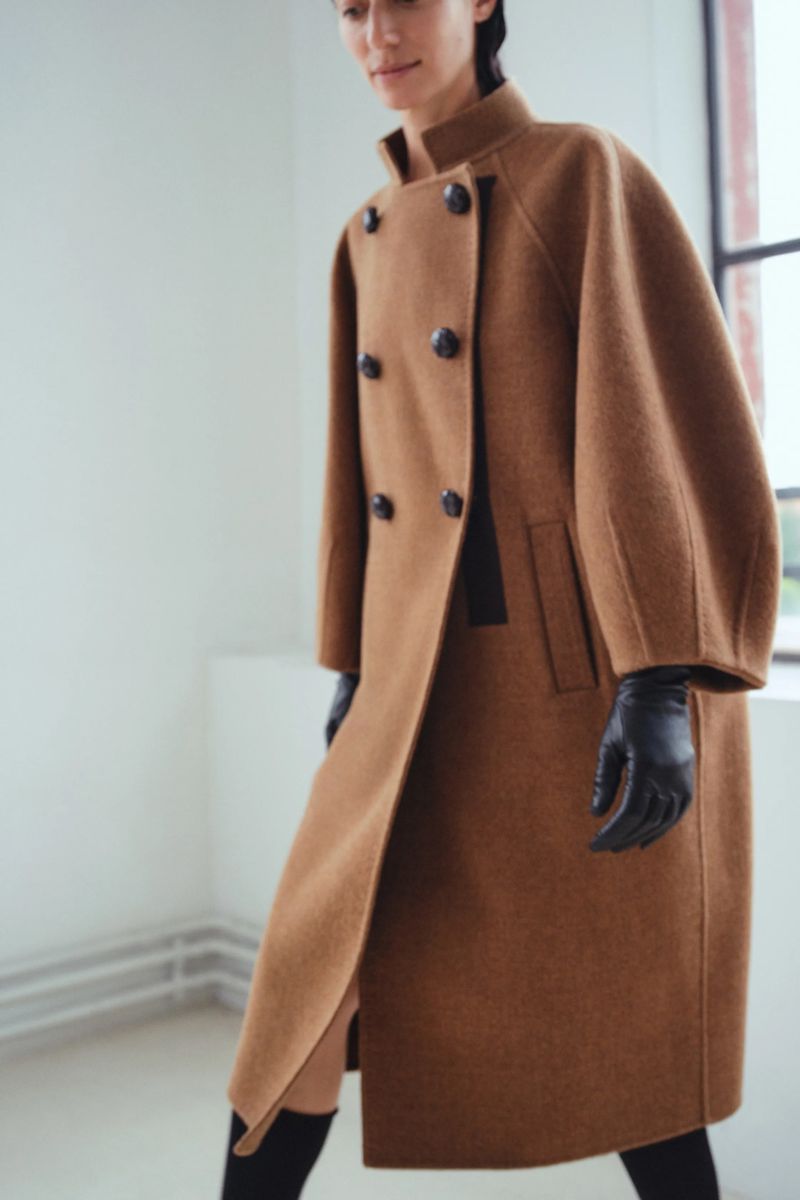 A woman models a double-breasted brown overcoat with black gloves inside a brightly lit indoor space near a window.