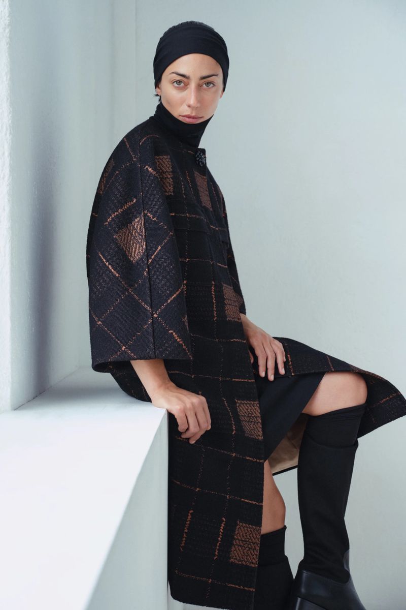 Model wearing a black grid-patterned coat, black headwrap, and tall black boots seated against a white studio wall.