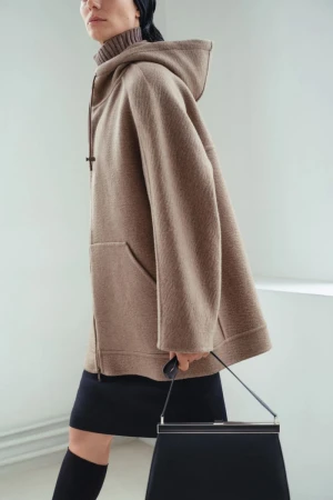 Fashionable woman in a hooded coat and knee-high socks carries a black handbag against a bright white background indoors.