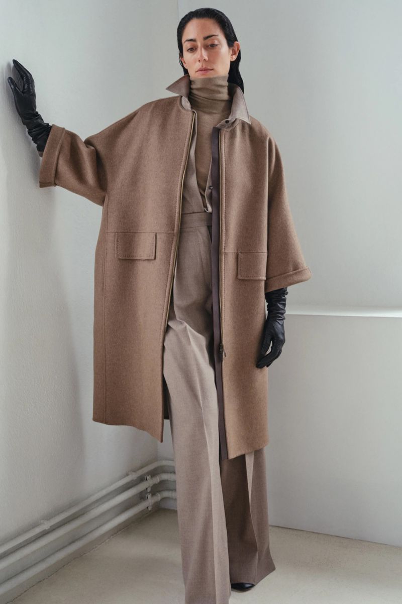 Fashionable woman poses in a studio wearing a brown wool coat, turtleneck, and wide-legged trousers. She accessorizes with long black gloves.