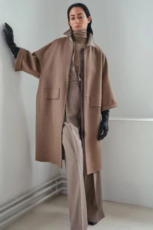Fashionable woman poses in a studio wearing a brown wool coat, turtleneck, and wide-legged trousers. She accessorizes with long black gloves.