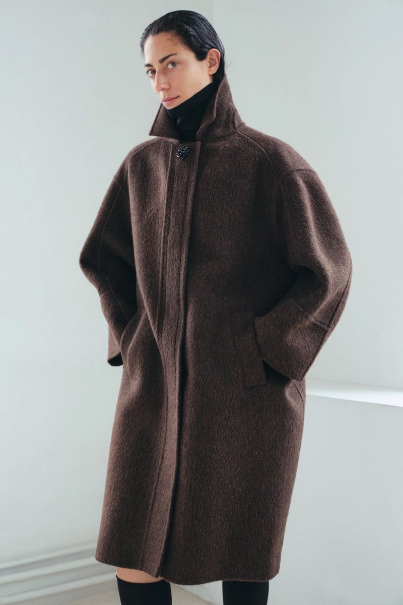 Woman in a stylish brown wool coat stands indoors in front of a white wall; a fashion-editorial studio shot.