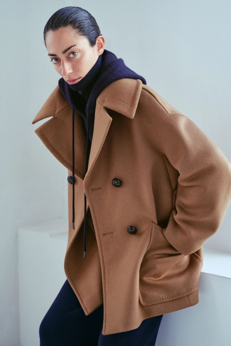 A woman poses in a studio wearing a tan double-breasted coat over a blue turtleneck and dark pants, with clean, minimalist styling.
