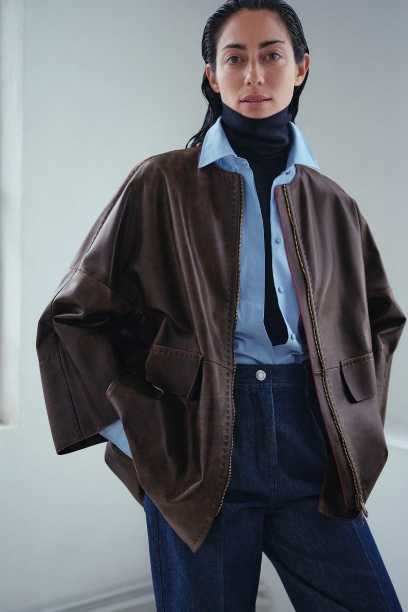 Adult female poses indoors wearing a brown leather jacket, light blue button-down shirt, black turtleneck, and dark blue denim pants.