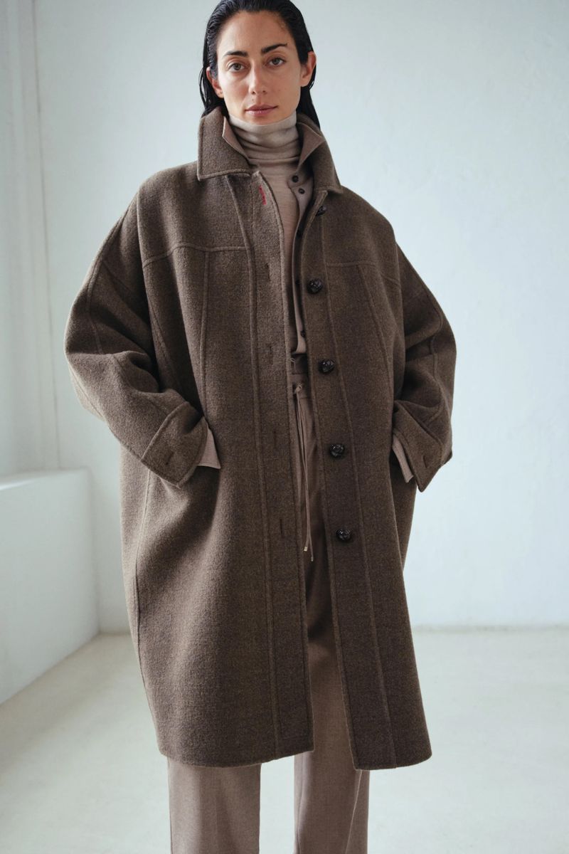 A woman models a brown wool overcoat, paired with a turtleneck and trousers, against a muted studio backdrop in a fashion editorial.