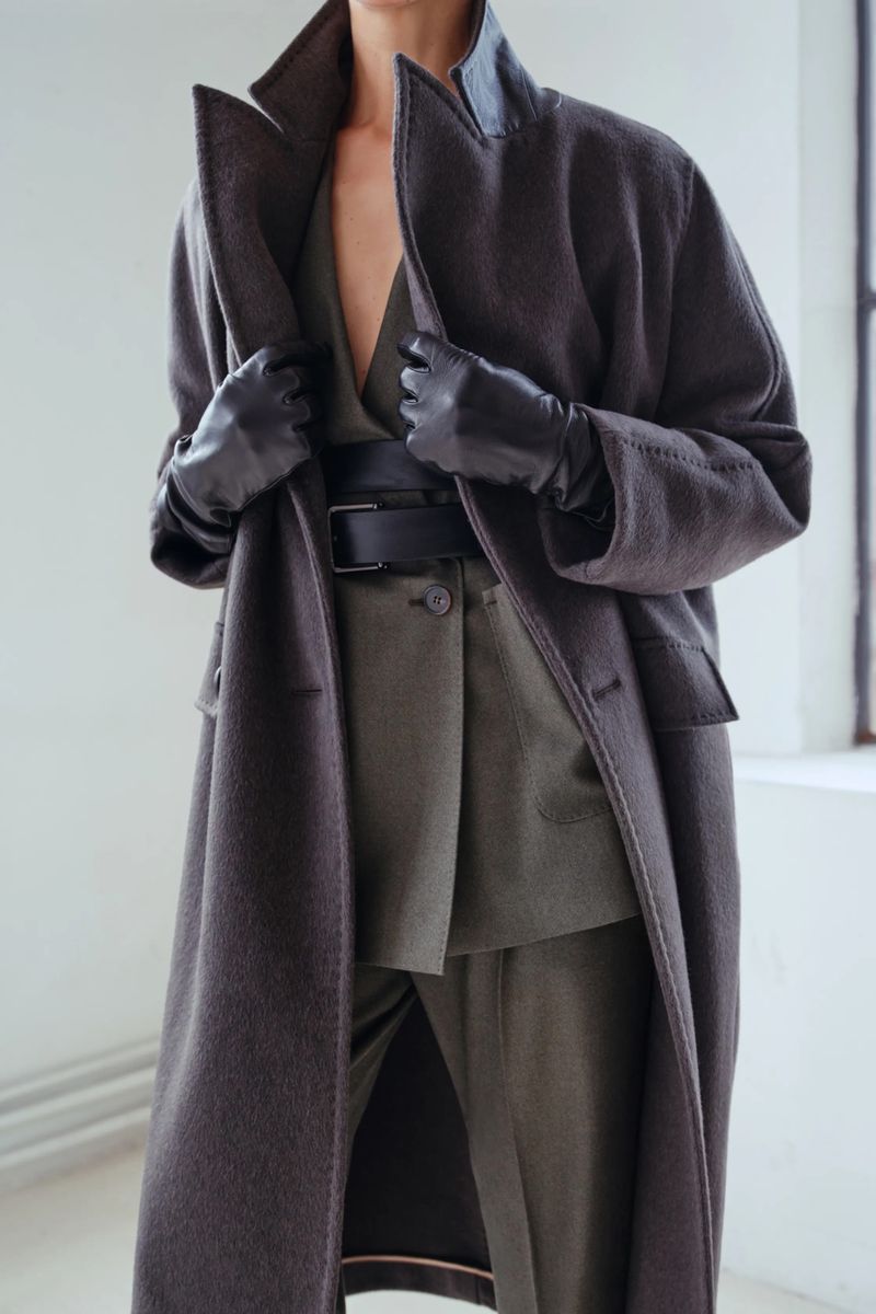 A woman poses indoors, wearing a long wool coat, gloves, and belt over a suit, standing near a window in a fashion-editorial style shot.