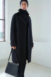 Model wearing a long black tailored coat and black headwrap, holding a black structured handbag in a bright white room.