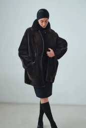 Model wearing an oversized dark brown plush shearling coat with a black head cap and skirt against a pale studio background.