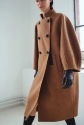 Model wearing a structured camel wool double-breasted coat with wide sleeves and black leather gloves in a bright studio interior.