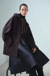 Model seated wearing a dark brown hooded zip jacket and structured black skirt for Max Mara Atelier Fall/Winter 2025.